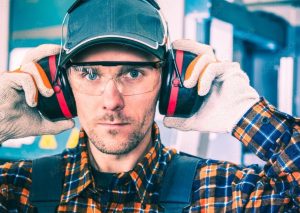 Worker in a checkered shirt and cap adjusts over-ear hearing protection while wearing safety glasses and gloves in an industrial setting.
